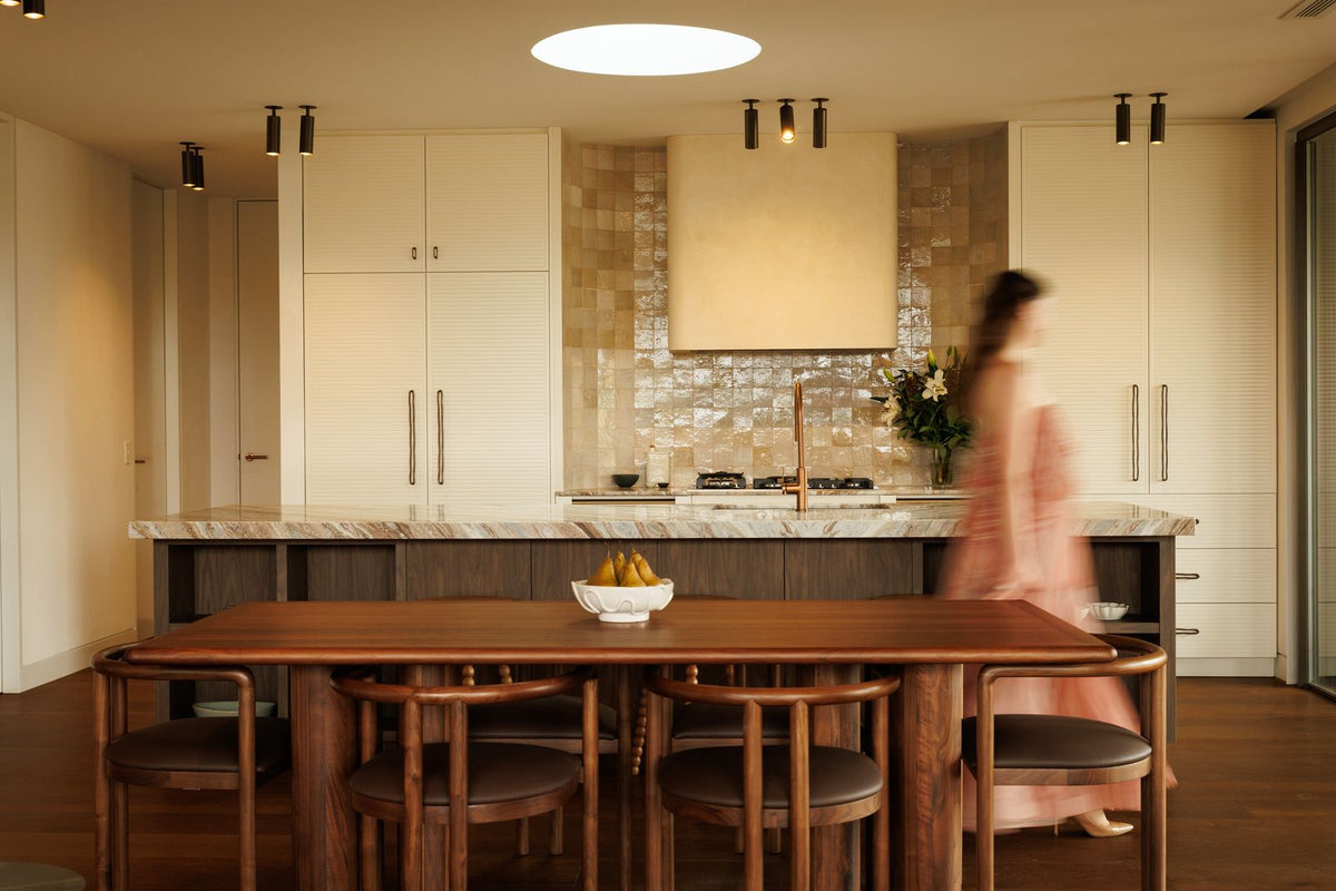 A modern kitchen interior with cream cabinets, a marble countertop, and a wooden dining table with chairs. A woman in a pink dress is blurred in motion near the counter.