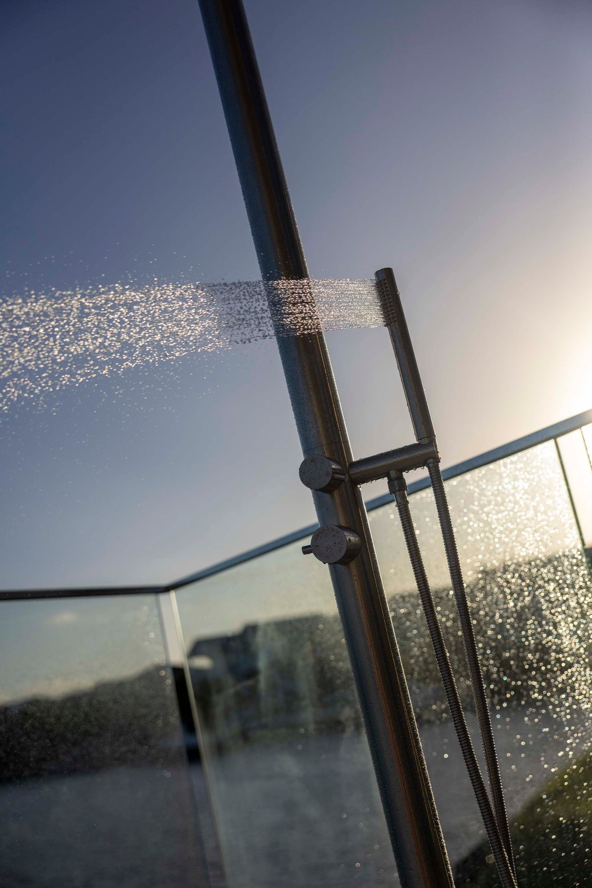 A close-up outdoor shower head spraying water onto a glass railing at sunset, with the water droplets illuminated by the warm sunlight.