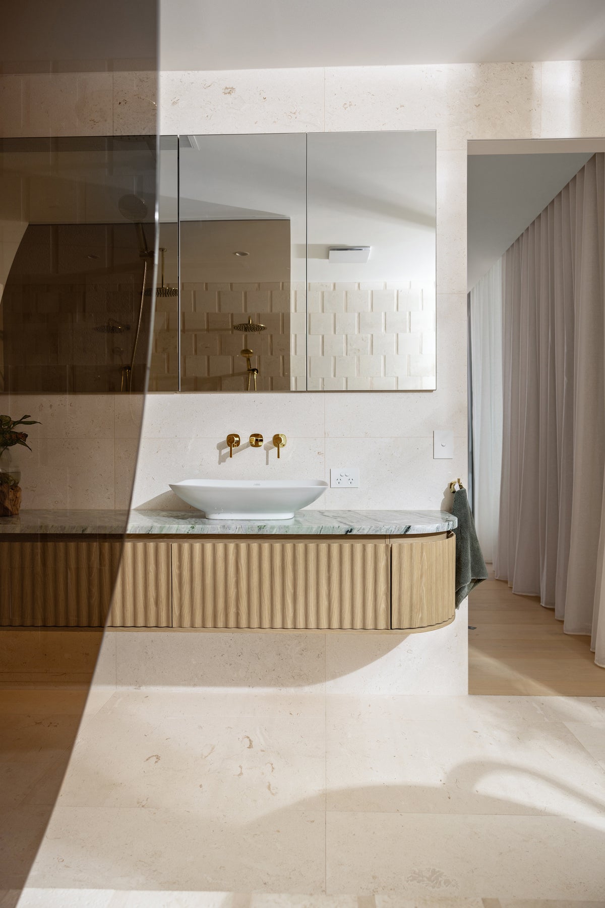 Modern bathroom vanity with a curved ribbed wood cabinet, a white vessel sink, a marble countertop, and a large wall mirror. The space features warm beige stone tile walls and floor, with soft natural light casting shadows across the foreground.